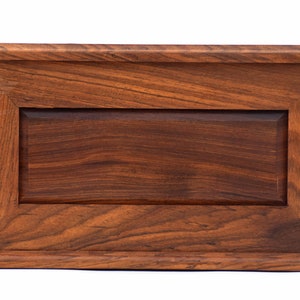 May include: A rectangular wooden box with a rich, dark brown grain pattern. The box features a raised frame design surrounding a recessed central panel, with a flat top and base molding that extends slightly beyond the main body of the box.
