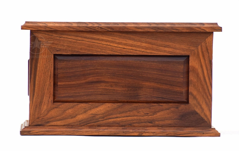 May include: A rectangular wooden box with a rich, dark brown grain pattern. The box features a raised frame design surrounding a recessed central panel, with a flat top and base molding that extends slightly beyond the main body of the box.