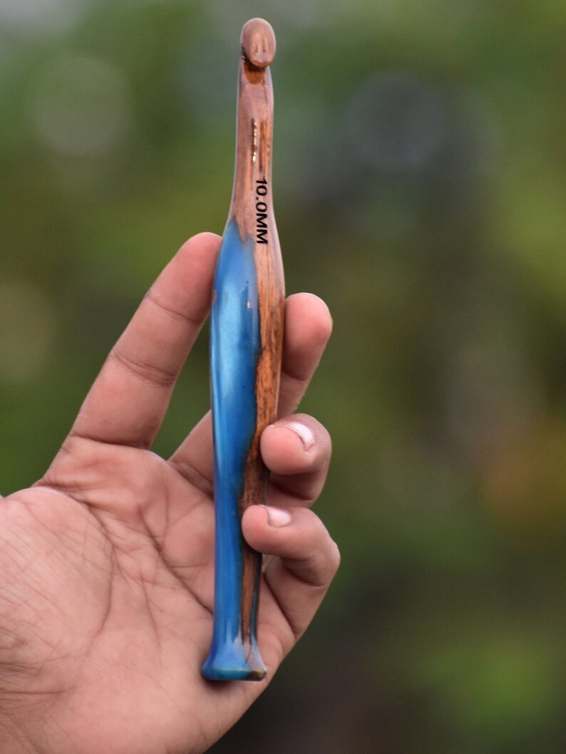 Pu&ograve; includere: Un uncinetto da 10,0 mm con un manico in legno levigato e una sezione in resina blu vibrante e traslucida. L'uncinetto presenta una testa affusolata e una base svasata, con la misura chiaramente stampata in nero sulla superficie in legno.