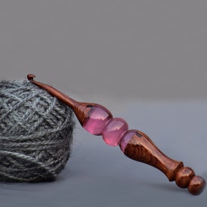 May include: A wooden crochet hook with a pink glass handle. The hook is lying on a gray surface next to a ball of gray yarn.