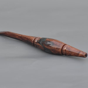 Puede incluir: Un ganchillo artesanal con un mango de madera de grano marr&oacute;n oscuro pulido y un acabado suave y brillante. La herramienta tiene una forma ergon&oacute;mica c&oacute;nica que se transforma en un gancho de metal delgado en la punta, mostrando vetas de madera natural.