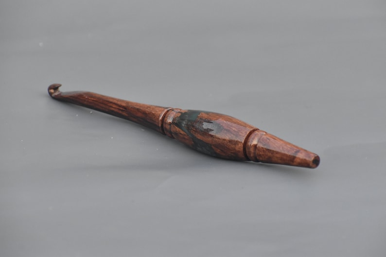 Puede incluir: Un ganchillo artesanal con un mango de madera de grano marr&oacute;n oscuro pulido y un acabado suave y brillante. La herramienta tiene una forma ergon&oacute;mica c&oacute;nica que se transforma en un gancho de metal delgado en la punta, mostrando vetas de madera natural.