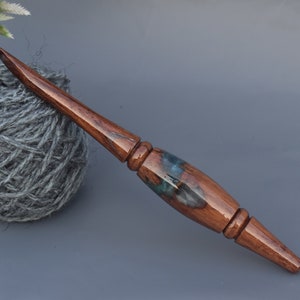Puede incluir: Un ganchillo artesanal con un eje de madera pulida de color marr&oacute;n oscuro y una secci&oacute;n central en el mango hecha de una combinaci&oacute;n de madera oscura y resina azul transl&uacute;cida con patrones de remolinos. El ganchillo tiene un dise&ntilde;o liso y c&oacute;nico.