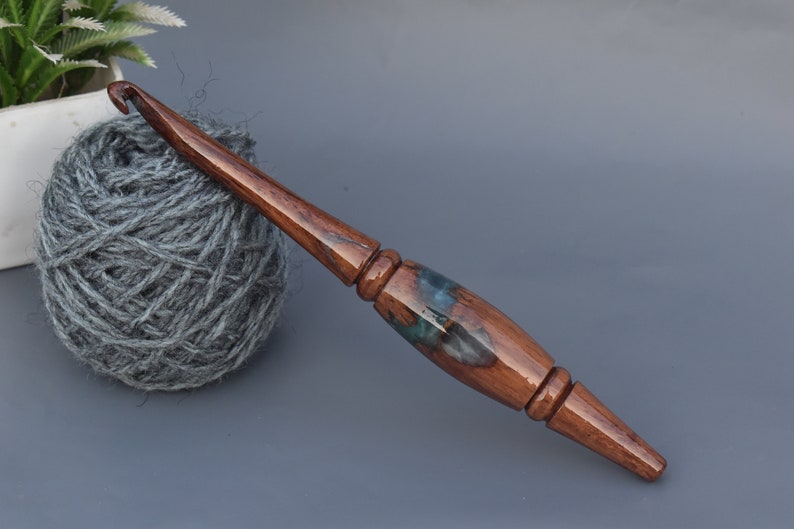 Puede incluir: Un ganchillo artesanal con un eje de madera pulida de color marr&oacute;n oscuro y una secci&oacute;n central en el mango hecha de una combinaci&oacute;n de madera oscura y resina azul transl&uacute;cida con patrones de remolinos. El ganchillo tiene un dise&ntilde;o liso y c&oacute;nico.
