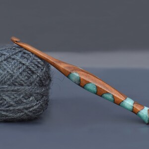 K&ouml;nnte beinhalten: Ein H&auml;kelnadel aus Holz mit einem teal- und braunen Marmormuster. Die Nadel liegt auf einer grauen Oberfl&auml;che neben einem Kn&auml;uel grauer Wolle.