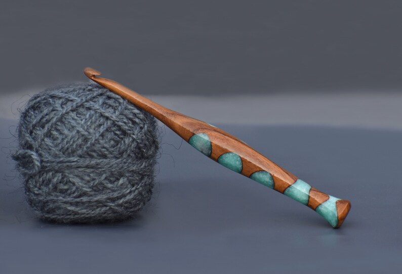 K&ouml;nnte beinhalten: Ein H&auml;kelnadel aus Holz mit einem teal- und braunen Marmormuster. Die Nadel liegt auf einer grauen Oberfl&auml;che neben einem Kn&auml;uel grauer Wolle.