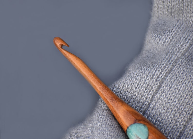 K&ouml;nnte beinhalten: Ein H&auml;kelnadel aus Holz mit einer t&uuml;rkisfarbenen und braunen Einlage am Ende. Die Nadel liegt auf einem grauen Strickstoff.