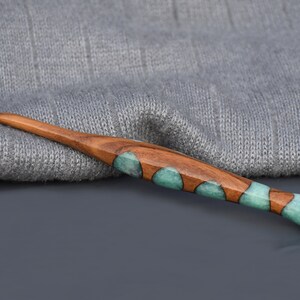K&ouml;nnte beinhalten: Ein H&auml;kelnadel aus Holz mit einem teal- und braunen Marmormuster. Die Nadel liegt auf einem grauen Strickstoff.