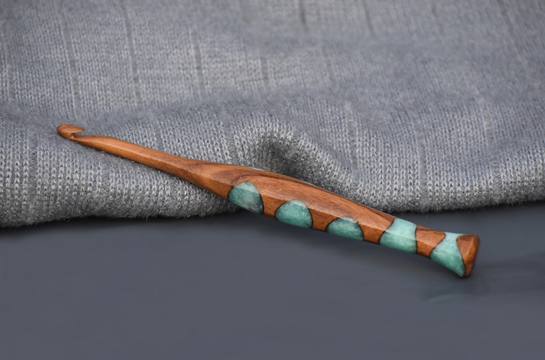 K&ouml;nnte beinhalten: Ein H&auml;kelnadel aus Holz mit einem teal- und braunen Marmormuster. Die Nadel liegt auf einem grauen Strickstoff.