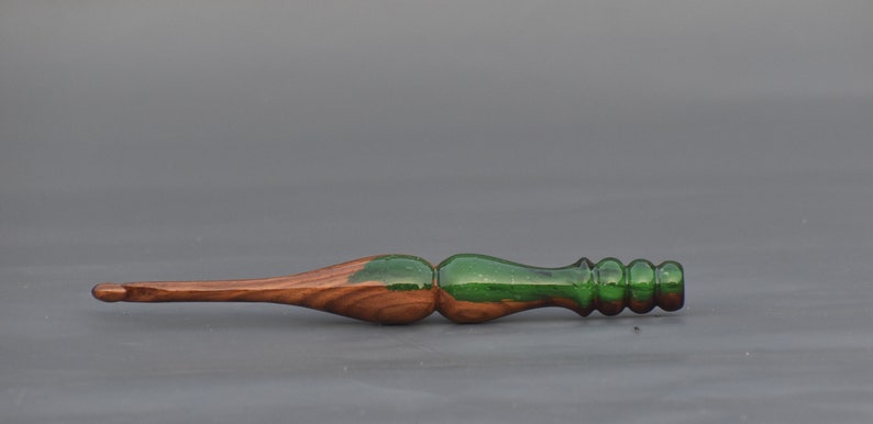 K&ouml;nnte beinhalten: Ein H&auml;kelnadel aus Holz mit einem gr&uuml;nen Harzgrif. Die Nadel ist spitz zulaufend und hat eine glatte Oberfl&auml;che.