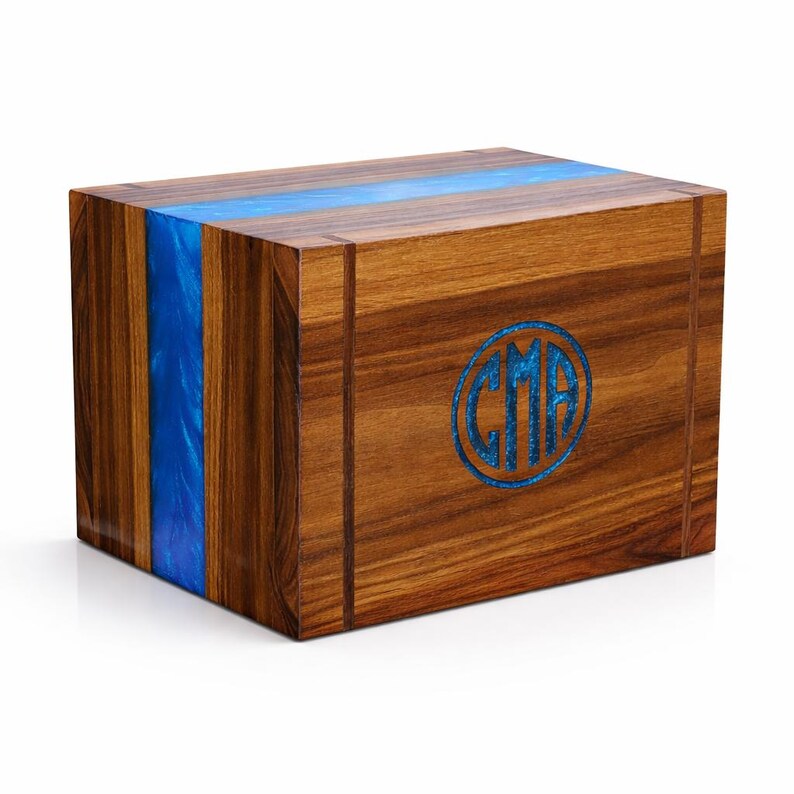 May include: A wooden box with a rich brown finish and a vibrant blue resin stripe and inlay. The box features a circular blue inlay with the letters "CMA". The design combines natural wood grain with a modern aesthetic.