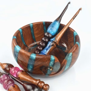 Pu&ograve; includere: Una ciotola rotonda in legno per filati con inserti verticali in resina turchese, accompagnata da diversi uncinetti. Gli uncinetti hanno manici in legno scuro con accenti in resina traslucida blu, rosa e viola glitterato, ciascuno contrassegnato da 6,00 mm.