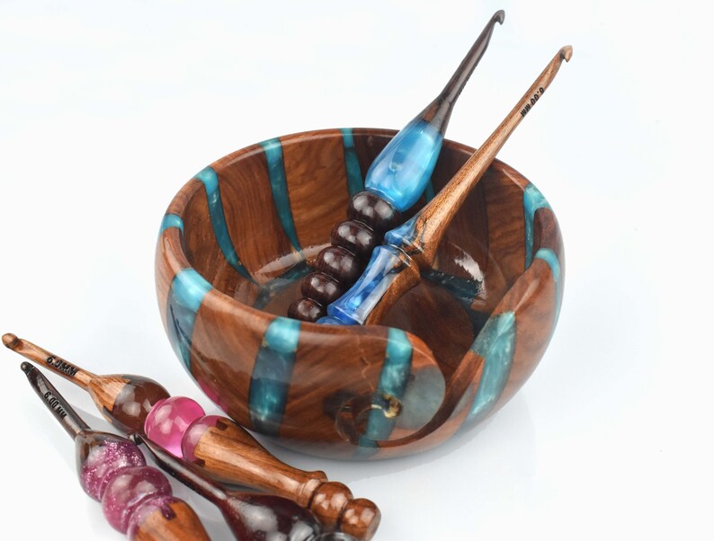 Pu&ograve; includere: Una ciotola rotonda in legno per filati con inserti verticali in resina turchese, accompagnata da diversi uncinetti. Gli uncinetti hanno manici in legno scuro con accenti in resina traslucida blu, rosa e viola glitterato, ciascuno contrassegnato da 6,00 mm.