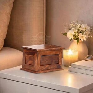 May include: A rectangular wooden box with a rich brown grain pattern and a smooth, polished finish. The box features a flat lid and a structured, paneled design on its sides, showcasing the natural wood texture and deep, warm color tones throughout.