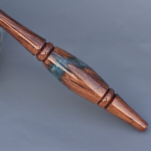 Puede incluir: Un ganchillo de madera pulida con un mango liso y c&oacute;nico que presenta una incrustaci&oacute;n decorativa central de resina azul oscuro y verde azulado. La madera tiene un tono marr&oacute;n c&aacute;lido y rico con vetas visibles y un acabado brillante.