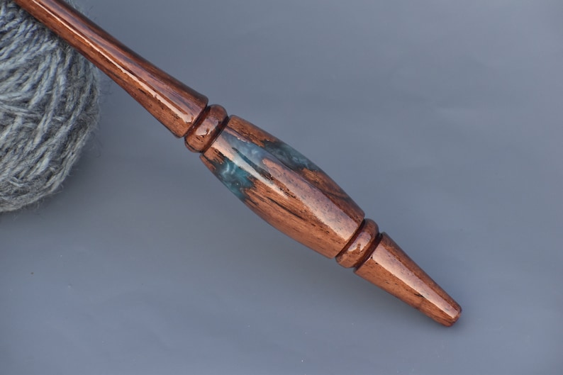 Puede incluir: Un ganchillo de madera pulida con un mango liso y c&oacute;nico que presenta una incrustaci&oacute;n decorativa central de resina azul oscuro y verde azulado. La madera tiene un tono marr&oacute;n c&aacute;lido y rico con vetas visibles y un acabado brillante.