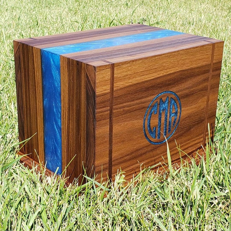 May include: A wooden box with a blue resin stripe and a blue CMA monogram. The box is made of dark wood and sits on a grassy surface. The blue resin adds a decorative element to the box.