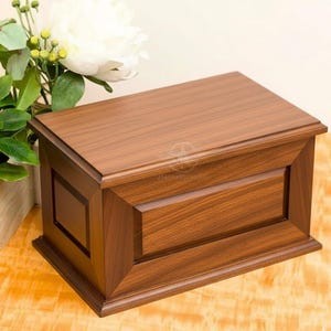 May include: A rectangular wooden box with a medium brown finish and a visible wood grain pattern. The box features a flat lid, a beveled frame design on the front panel, and a raised base. The construction includes clean, straight edges.