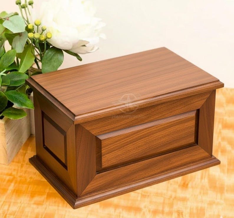 May include: A rectangular wooden box with a medium brown finish and a visible wood grain pattern. The box features a flat lid, a beveled frame design on the front panel, and a raised base. The construction includes clean, straight edges.