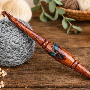 Puede incluir: Un ganchillo artesanal con mango de madera pulida en color marr&oacute;n oscuro y una incrustaci&oacute;n central de resina con remolinos azules y negros. La herramienta presenta un dise&ntilde;o liso y c&oacute;nico con un gancho met&aacute;lico curvado en la punta.