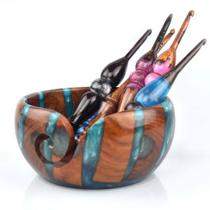 Pu&ograve; includere: Una ciotola rotonda in legno per filati con intarsi verticali in resina color ottanio e un'apertura a spirale, contenente quattro uncinetti artigianali con manici in legno levigato e dettagli in resina nei toni del nero, rosa e blu.