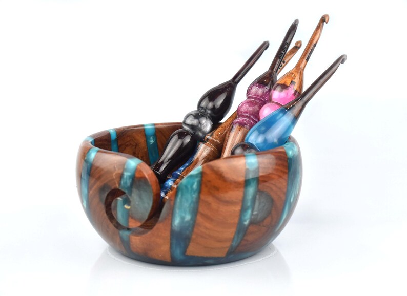 Pu&ograve; includere: Una ciotola rotonda in legno per filati con intarsi verticali in resina color ottanio e un'apertura a spirale, contenente quattro uncinetti artigianali con manici in legno levigato e dettagli in resina nei toni del nero, rosa e blu.