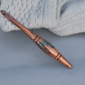 Puede incluir: Un ganchillo artesanal con mango de madera pulida en color marr&oacute;n oscuro y una secci&oacute;n central de resina verde oscuro transl&uacute;cida. La herramienta presenta una forma ergon&oacute;mica y c&oacute;nica con un acabado liso y una punta met&aacute;lica en el extremo.