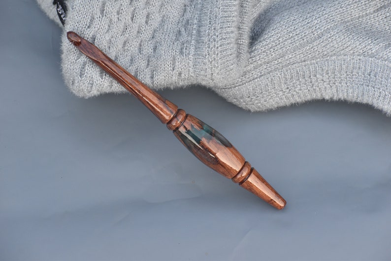 Puede incluir: Un ganchillo artesanal con mango de madera pulida en color marr&oacute;n oscuro y una secci&oacute;n central de resina verde oscuro transl&uacute;cida. La herramienta presenta una forma ergon&oacute;mica y c&oacute;nica con un acabado liso y una punta met&aacute;lica en el extremo.