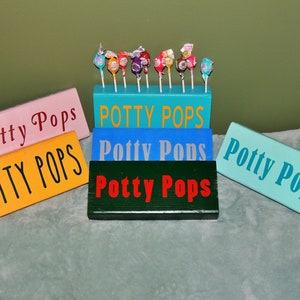 May include: Five wooden signs with the text "Potty Pops" in different colors and fonts. The signs are arranged in a staggered pattern, with the top sign being blue and the bottom sign being turquoise. The signs are decorated with lollipops.