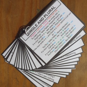 May include: A set of flashcards with black borders, held together by a silver ring. The top card displays "SINGLE AND PLURAL" with grammar rules. Examples include "Dog/Dogs" and "Toy/Toys." The cards are fanned out on a wooden surface.