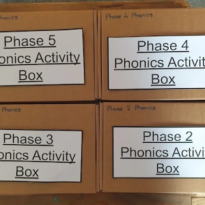 May include: Four cardboard boxes labelled with white paper and black text. The boxes are labelled "Phase 5 Phonics Activity Box", "Phase 4 Phonics Activity Box", "Phase 3 Phonics Activity Box", and "Phase 2 Phonics Activity Box".