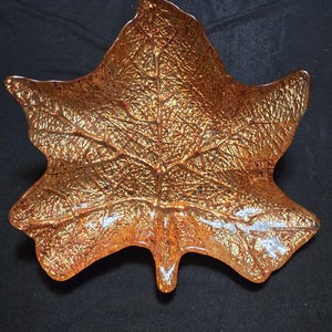 May include: A decorative, amber-colored glass leaf-shaped dish. The dish has a textured surface with detailed vein patterns. The leaf dish is a unique accent piece for home decor, measuring approximately 10 inches in length.