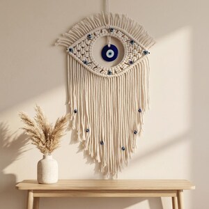 May include: A wall hanging made of off-white macrame cord featuring an eye-shaped design. The center contains a circular blue and white glass evil eye charm, with smaller matching charms integrated into the knotted cord and long fringe.
