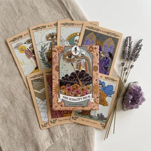 May include: A deck of tarot cards titled The Audacity Deck by The Pine Torch, featuring illustrated cards with skeletons, celestial motifs, and floral patterns, accompanied by a sprig of dried lavender and a purple amethyst crystal cluster.