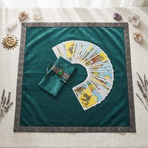 Peut inclure: Un carré de tissu en velours vert foncé avec une bordure décorative sombre, accompagné d'une pochette à cordon assortie ornée d'une pierre précieuse verte et d'un éventail de cartes de tarot, dont la carte intitulée The Fool.
