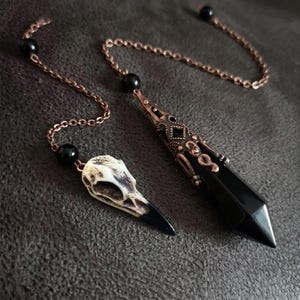 May include: Two pendulums on copper-toned chains with black beads. One pendulum features a polished black stone point with intricate copper-toned metal filigree. The other pendulum is a small, off-white bird skull with a dark, pointed beak.