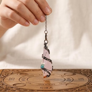May include: A faceted, pale pink crystal pendulum wrapped in dark, twisted metal wire. The wire features small, colourful accent beads in blue, white, and orange. The crystal hangs from a dark metal chain, suspended above a circular wooden board.