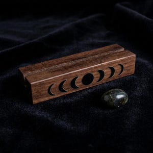 May include: A rectangular dark brown wooden stand featuring a carved moon phase cycle along the front side. The stand has a horizontal slot running across the top surface. A single smooth, dark, iridescent polished stone sits nearby.