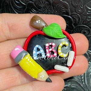 May include: A clay apple-shaped chalkboard ornament featuring a red border, a brown stem, and a green leaf. The black center displays the letters ABC in blue, pink, and yellow with white dots. A yellow pencil with a pink eraser is attached.