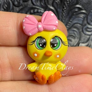 May include: A yellow chick figurine with large green eyes, orange feet, and a pink bow with white polka dots on its head. The chick has white dots on its cheeks and the text Dream Time Clays is printed across the lower portion of the figure.