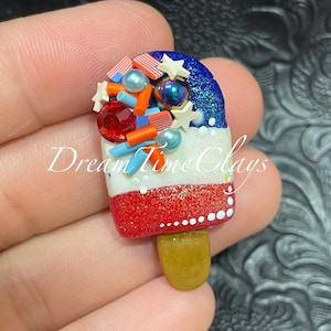 May include: A red, white, and blue popsicle with a brown stick. The popsicle is covered in sprinkles and stars. The popsicle is made of polymer clay.