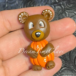May include: A brown bear figurine holding a bright orange pumpkin. The bear has white dots on its ears, large eyes, and a black nose. The text Dream Time Clays is written across the center of the bear and pumpkin. The bear has brown paws and feet.