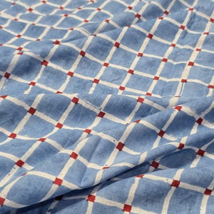 Op de afbeelding: Een lichtblauwe stof met een wit ruitpatroon en kleine rode vierkantjes op elk kruispunt. Het materiaal heeft een zachte, geplooide textuur en toont een herhalend geometrisch ontwerp over het gehele oppervlak van het textiel.