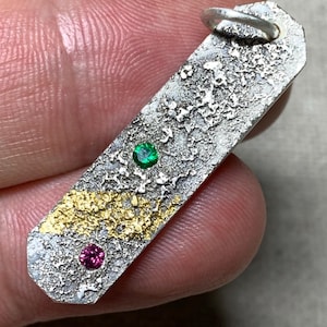 Könnte beinhalten: Ein rechteckiger Silberanhänger mit einer stark texturierten, oxidierten Oberfläche und einem Bereich aus Blattgold. Der Anhänger ist mit einem kleinen runden grünen Edelstein und einem kleinen runden rosa Edelstein besetzt und hat eine Silberöse.