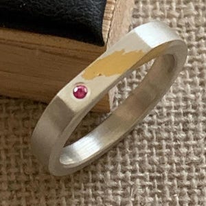 Peut inclure: Une bague anneau argentée avec une finition mate brossée, une petite pierre précieuse rose circulaire sertie dans le métal et une tache dorée irrégulière sur la surface supérieure de l'anneau, créant un contraste marqué de texture et de couleur.