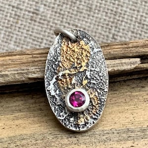 May include: An oval-shaped pendant featuring a textured, oxidized silver surface with patches of gold-toned metal. A single, round, vibrant pink gemstone is bezel-set in the lower portion of the pendant, which includes a small silver jump ring at the top.