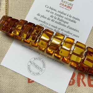 May include: A close-up of a golden amber bracelet with a square design. The bracelet is on a white card with the text "Nature d'Ambre" and "Ce bijou, réalisé à la main, est en Ambre véritable de plus de 40 millions d'années. de la Baltique This jewellery is made of 40 million years old Genuine Baltic Amber or amber and sterling silver item of jewellery Baltic Amber Authentic Baltic Amber".
