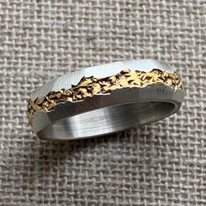 Op de afbeelding: Een zilverkleurige ring met een getextureerde, grillige band en een centrale inleg van ruw, goudkleurig metaalachtig materiaal. Het buitenoppervlak van de ring heeft een geborstelde afwerking, wat contrasteert met het onregelmatige, kristallijne uiterlijk van het goudkleurige middengedeelte dat horizontaal rond de omtrek loopt.