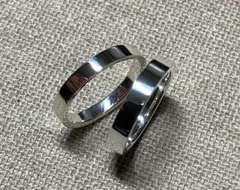 Könnte beinhalten: Zwei glänzende Silberringe mit flachem Banddesign. Die Ringe werden auf einer strukturierten, neutralfarbenen Oberfläche gezeigt. Die Ringe reflektieren das Licht und erzeugen ein helles, poliertes Aussehen. Diese Ringe sind wahrscheinlich für eine Hochzeit oder Verlobung.