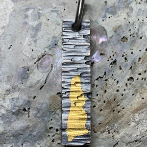 May include: A rectangular metal pendant featuring a dark, oxidized, and heavily textured surface with a vertical, irregular patch of bright gold-colored inlay. A small, smooth, silver-toned metal jump ring is attached to the top of the pendant.